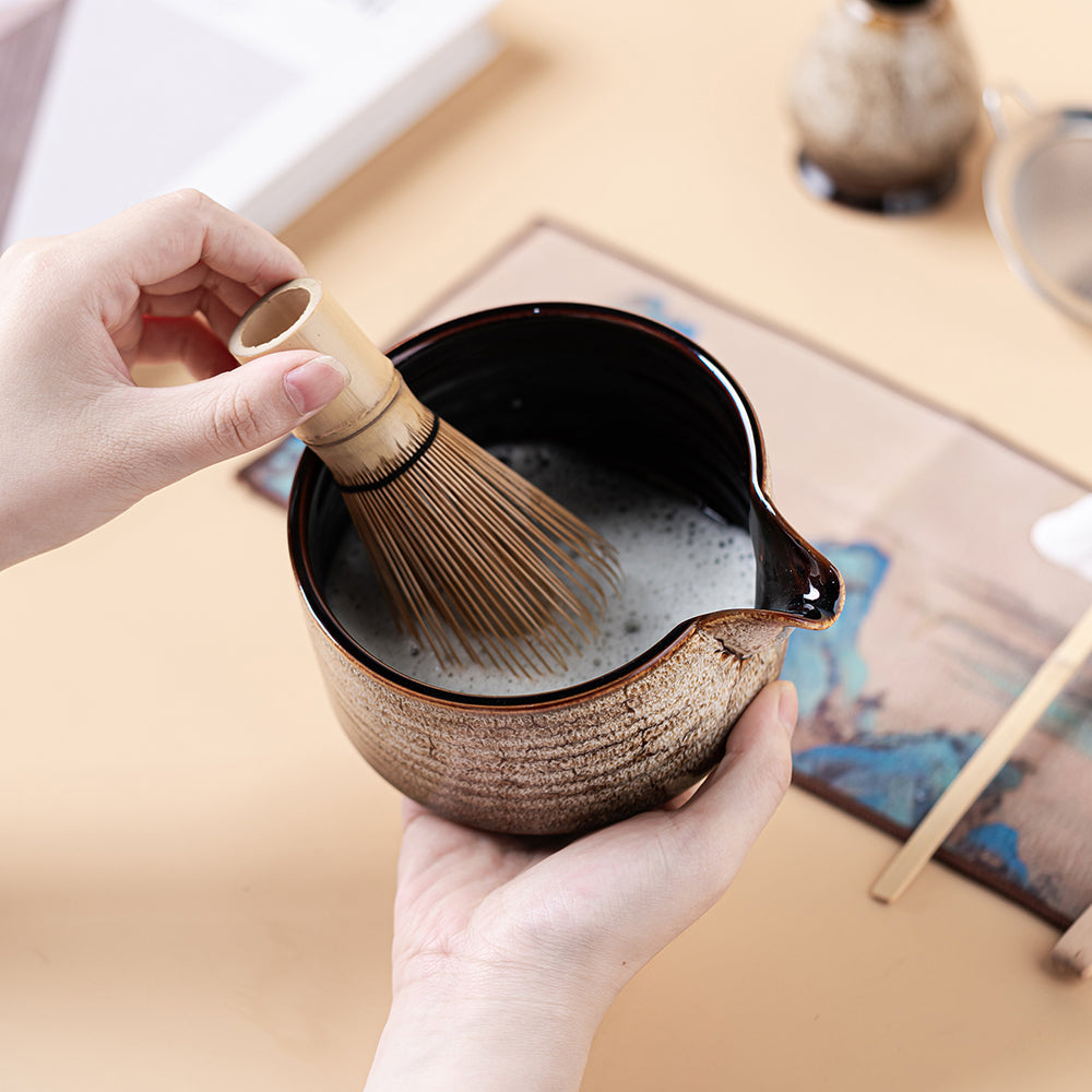 Hand holding bamboo whisk over ceramic tea set for matcha